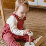 Cute baby in overalls playing with a wooden toy on a carpeted floor indoors.
